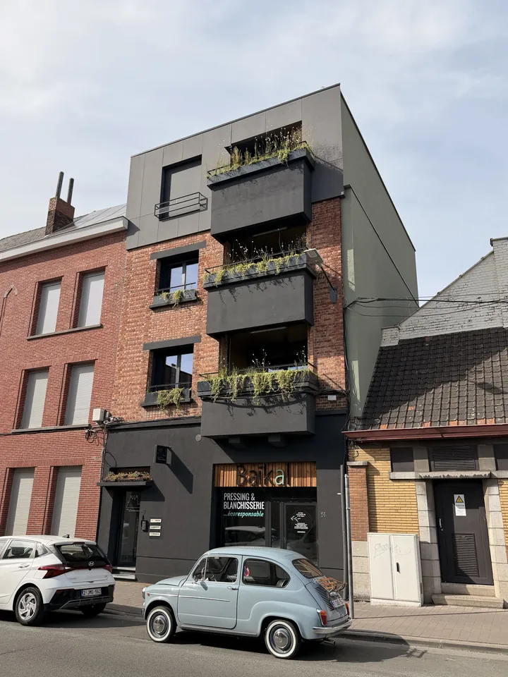 La façade de La Fonderie, boulevard Eisenhower 51 à Tournai : briques, balcons plantés, pressing au rez-de-chaussée et petite Fiat ancienne bleu ciel