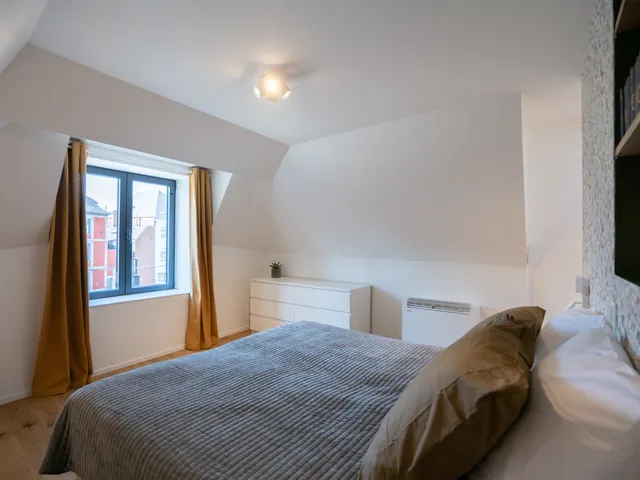 Chambre sous combles avec lit double, commode blanche, papier peint à motifs et fenêtre avec rideaux ocre, Camuche de Bettignies à Tournai