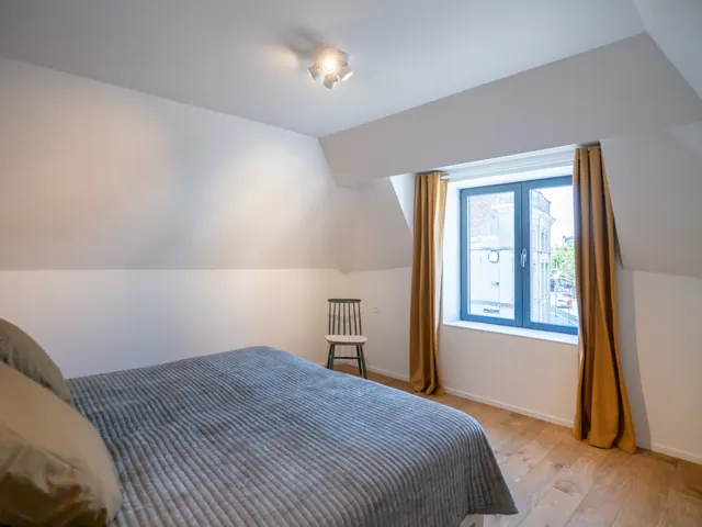 Chambre sous combles avec lit double, couvre-lit gris côtelé, rideaux ocre et fenêtre sur la ville, Camuche de Bettignies à Tournai