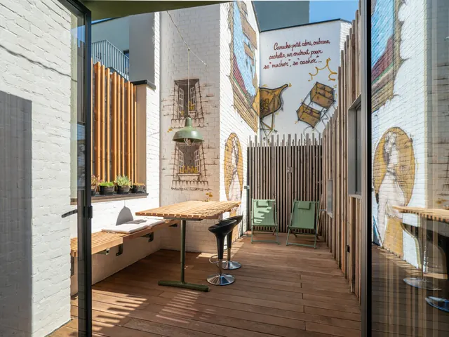 Terrasse en bois avec table à lattes, tabourets, transats verts et fresque murale qui définit le mot « camuche », Camuche de Bettignies à Tournai