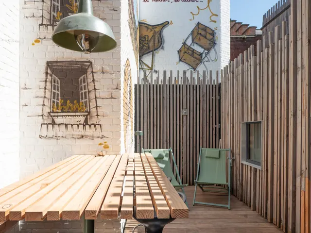 Terrasse avec grande table en bois à lattes et suspension verte, devant des fenêtres peintes sur un mur blanc, Camuche de Bettignies à Tournai
