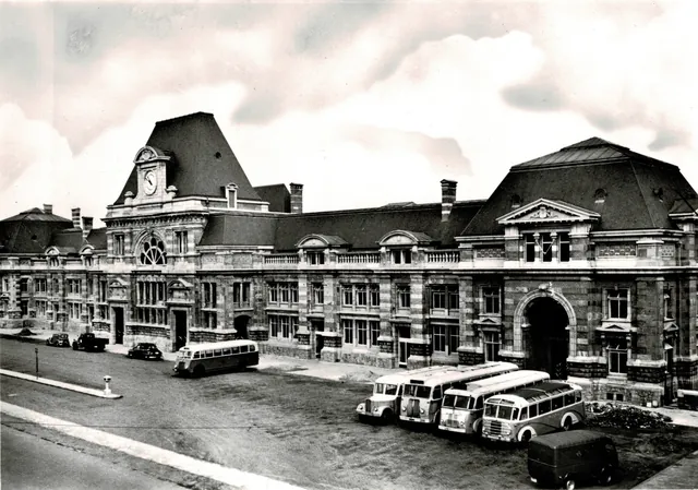 Gare de Tournai vue en légère plongée : façade à bandes de briques et de pierre, pavillon central à horloge, autocars stationnés sur la place, photographie ancienne en noir et blanc, archive