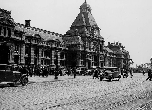 Façade de la gare de Tournai vue depuis la place pavée, voies de tramway au premier plan, automobiles anciennes et passants, photographie ancienne en noir et blanc, archive