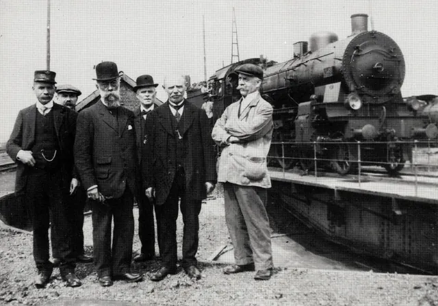 Six hommes en costume posant devant une locomotive à vapeur sur une plaque tournante, l'un porte une barbiche et un chapeau melon, reproduction imprimée d'une photographie ancienne, archive