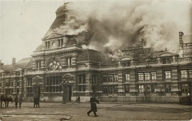 Carte postale ancienne : la gare de Tournai en flammes, fumée au-dessus de la toiture, pompiers et passants sur la place, archive