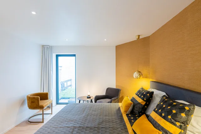 Chambre double : mur en tissu ocre, lit gris aux coussins jaunes, suspension dorée et fauteuil, Camuche Boulenger à Tournai