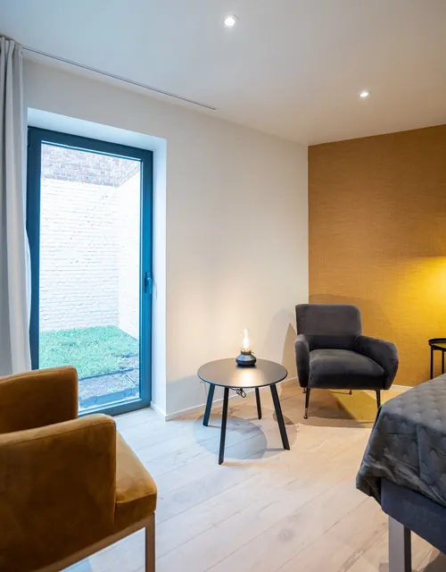 Coin détente de la chambre : fauteuil gris, table d'appoint et lampe devant la porte-fenêtre sur le jardin, Camuche Boulenger à Tournai