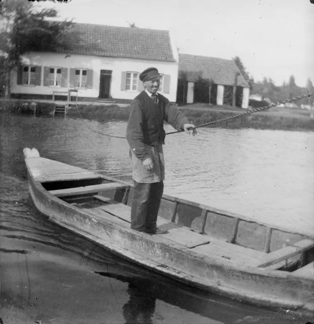 Homme debout dans un bac tirant sur une chaîne, devant une maison au bord de l'eau, photographie ancienne, Camuche Desclée à Tournai