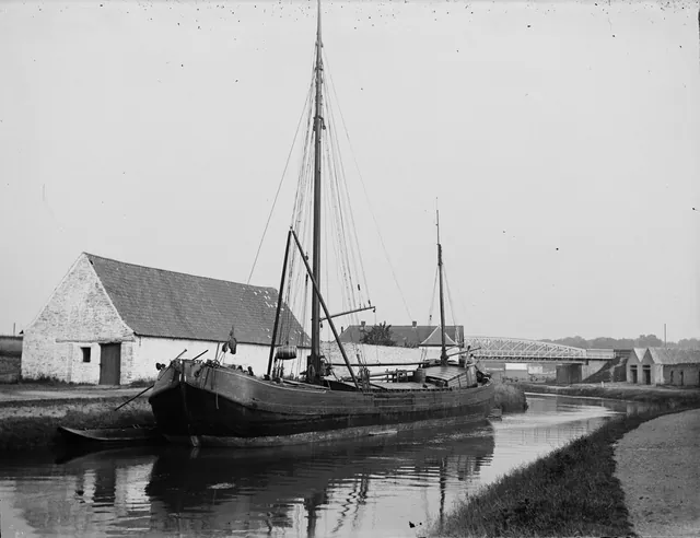 Péniche à voiles amarrée le long d'un canal, avec une grange en pierre et un pont métallique, photographie ancienne, Camuche Desclée à Tournai