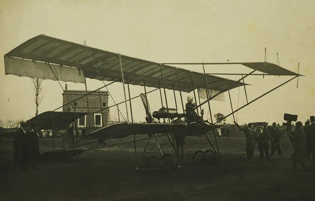 Biplan sur un terrain, pilote assise aux commandes, spectateurs et opérateur de cinéma à droite, photographie ancienne sépia, Camuche Dutrieu à Tournai