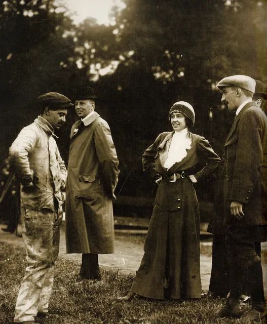 Hélène Dutrieu en bonnet de laine et jabot blanc, entourée de quatre hommes sur un terrain, photographie ancienne sépia redressée, Camuche Dutrieu à Tournai