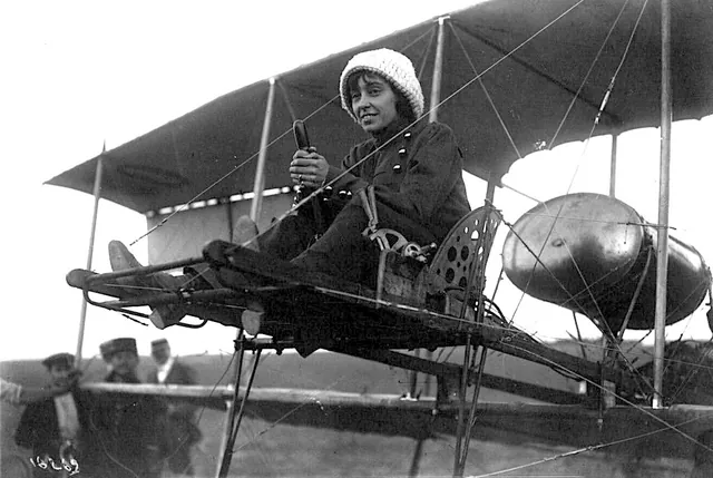 Hélène Dutrieu assise sur un biplan, la main sur la commande, des hommes en arrière-plan, photographie ancienne, Camuche Dutrieu à Tournai