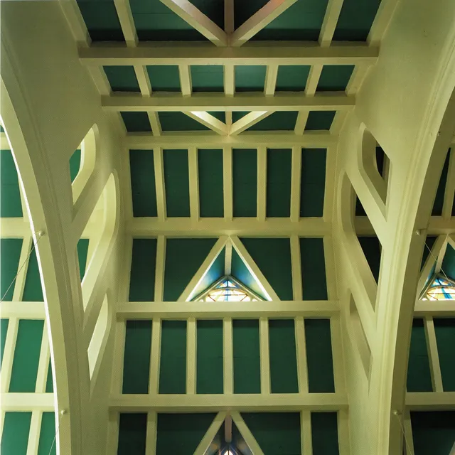 Vue plongeante sur une charpente de poutres crème peintes sur fond vert, piliers aux formes courbes et petits vitraux triangulaires, photographie, archive