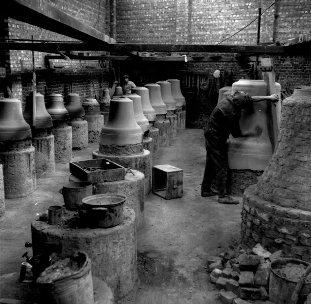 Atelier de la fonderie : rangées de cloches de bronze sur leurs socles de briques et d'argile, un ouvrier penché sur l'une d'elles, photographie ancienne, Camuche Michiels à Tournai