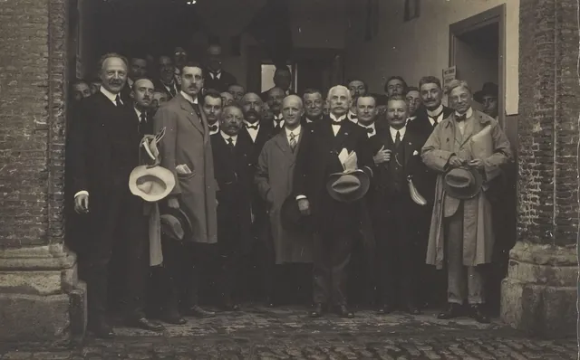 Groupe d'hommes en costume, chapeaux à la main, posant sous un porche de briques, tirage ancien à bords dentelés, Camuche Michiels à Tournai