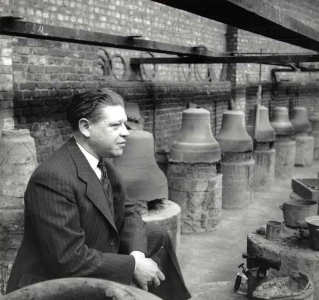 Marcel Michiels, en costume rayé, assis devant des rangées de moules et de cloches dans la cour de la fonderie, photographie ancienne, Camuche Michiels à Tournai